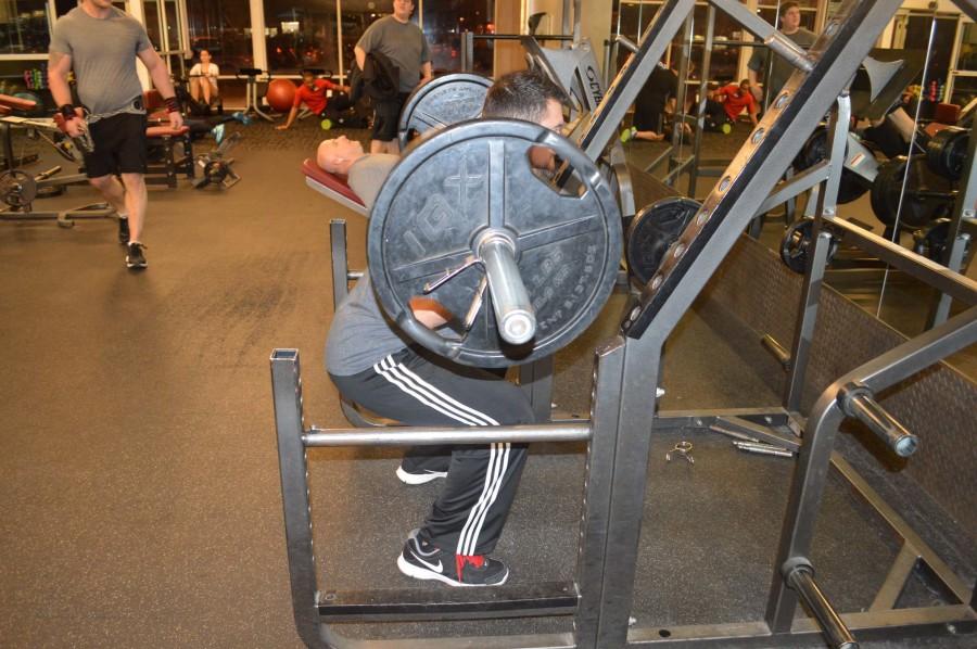 Campbell demonstrates the proper form of a squat by putting the bar behind his neck, resting it on his upper shoulders. He bends his knees until the back of his thigh and calf make a 90 degree angle, and then comes back up.  
