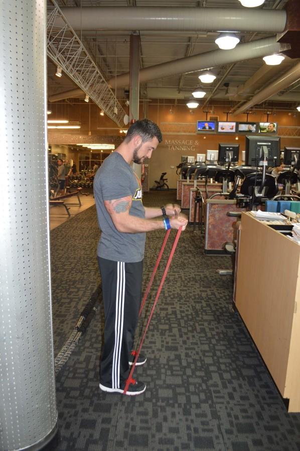 Campbell works his arms with a resistance band. While standing with his feet shoulder width apart, he holds the band down with his feet. He holds onto the top of the resistance band also shoulder width apart, and curls his arms up as far as possible. 
