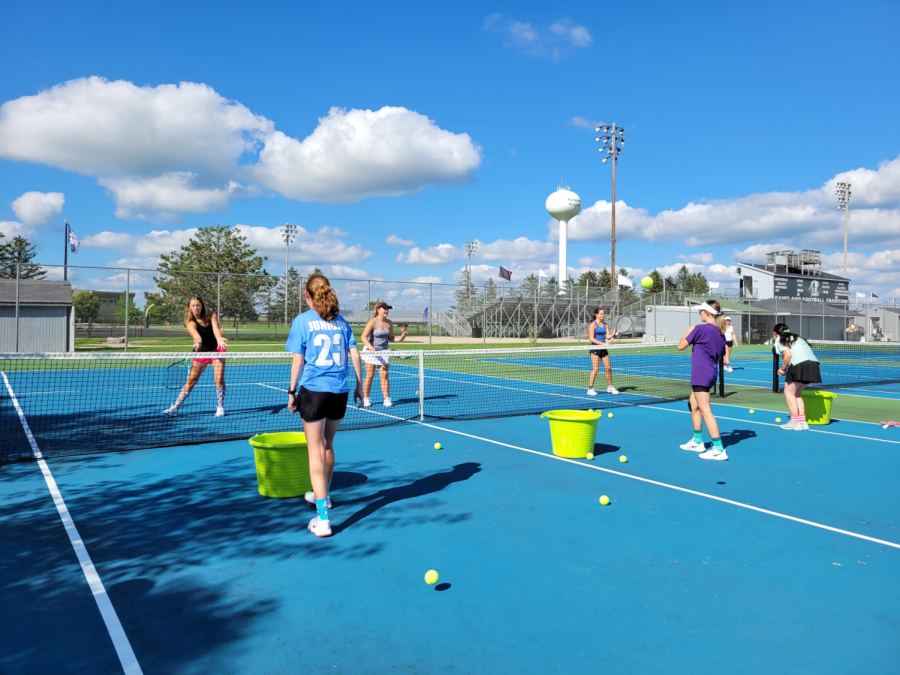 The varsity tennis team practices drills on the tennis courts. The team finds fun ways to complete repetitive tasks. 